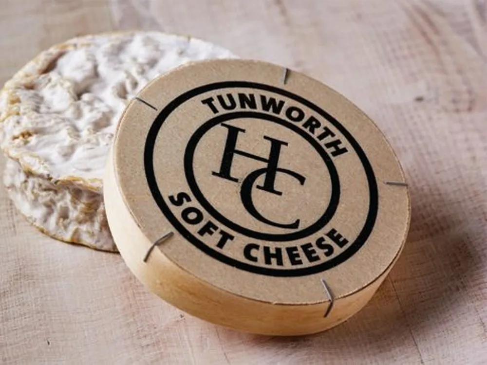 A disc of Tunworth cheese wrapped in paper sits next to its circular box, which displays its name