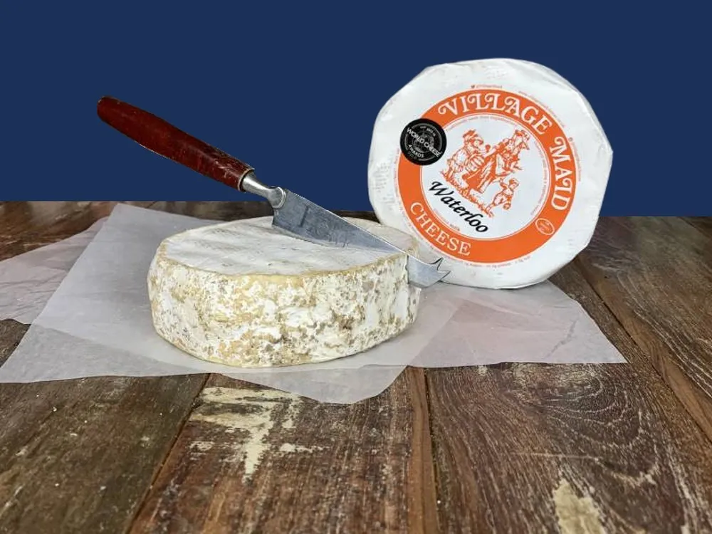 A disc-shaped Waterloo cheese is being sliced with a knife on a cutting board, next to a wheel of Waterloo cheese wrapped in white paper.