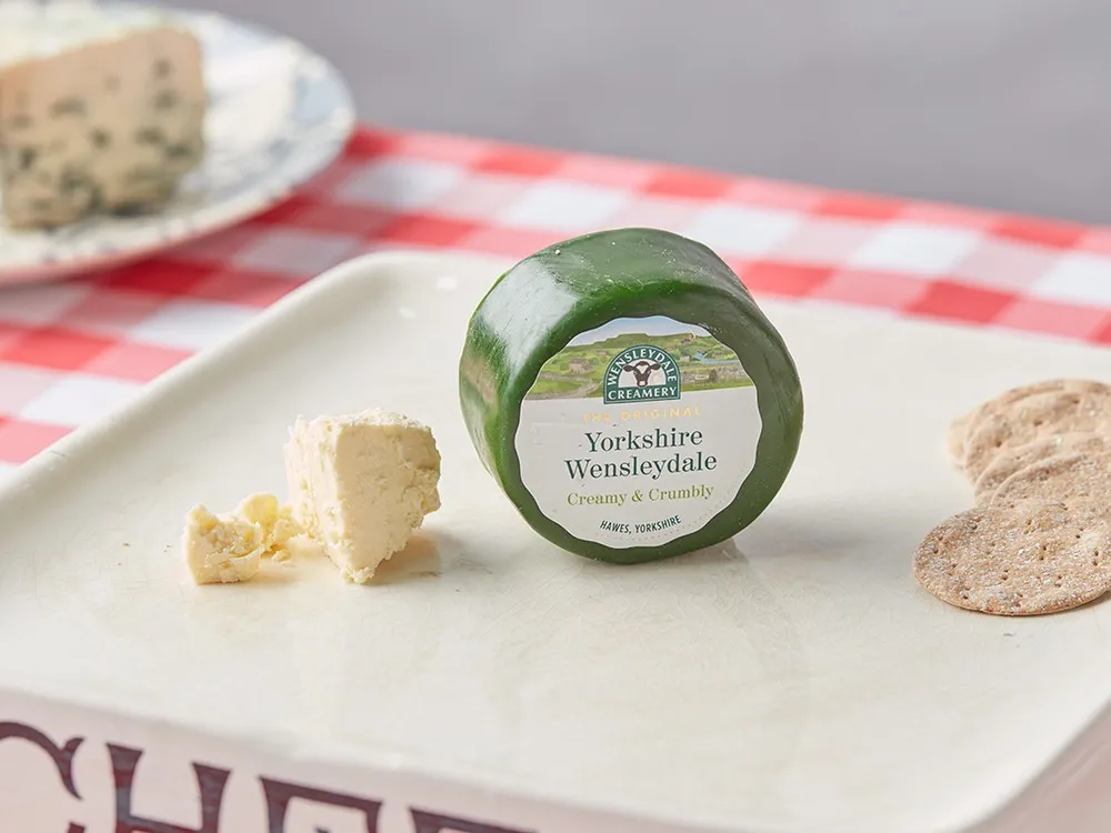 A Yorkshire Wensleydale cheese disc, wrapped in green wax, sits on a white plate alongside crackers, placed on a table