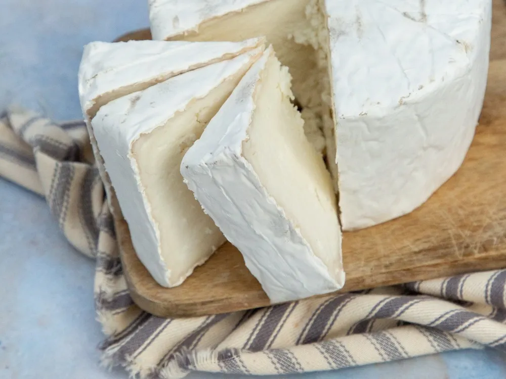 A round-shaped White Nancy cheese and slices showing creamy white bloomy rind sits on a wooden surface