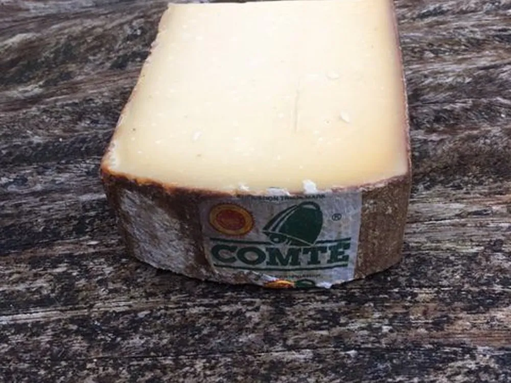 A slice of 'aged Comte 30 months' cheese showcasing its smoth, buttery texture rests on a wooden surface