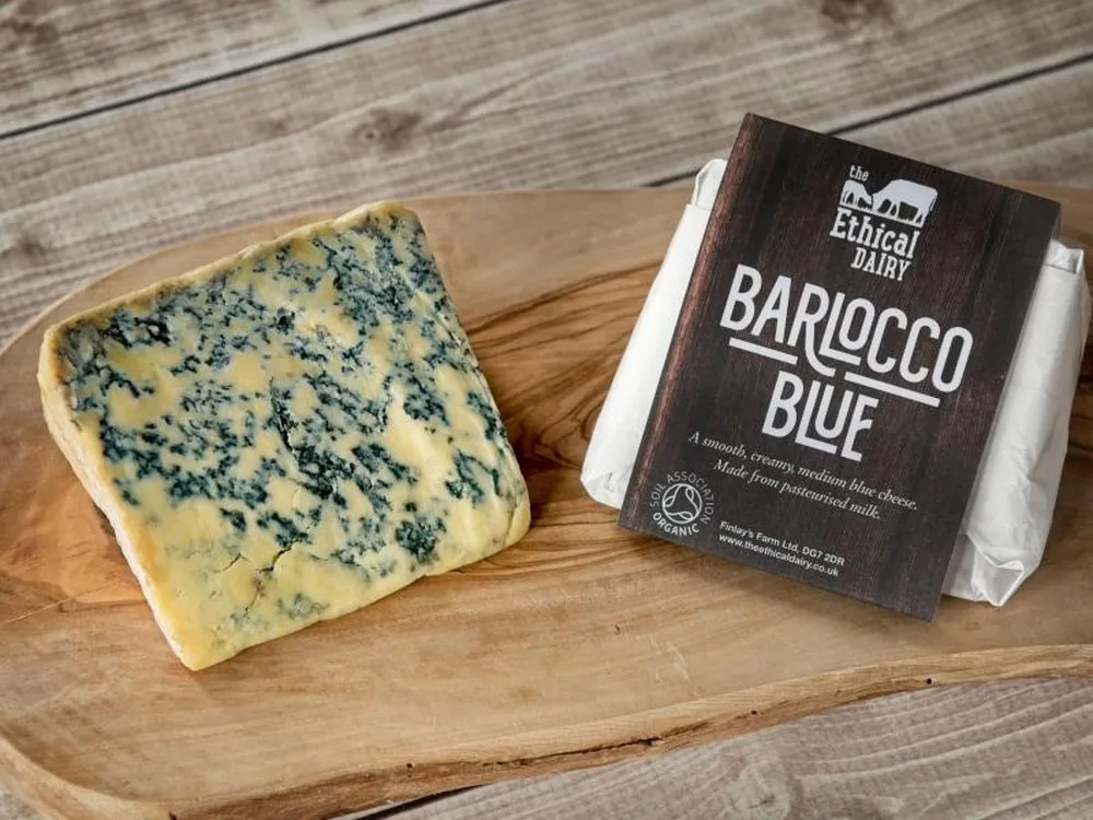 A piece of Blarlocco blue cheese, featuring prominent blue veins, sits on a wooden board beside another wrapped Blarlocco blue cheese