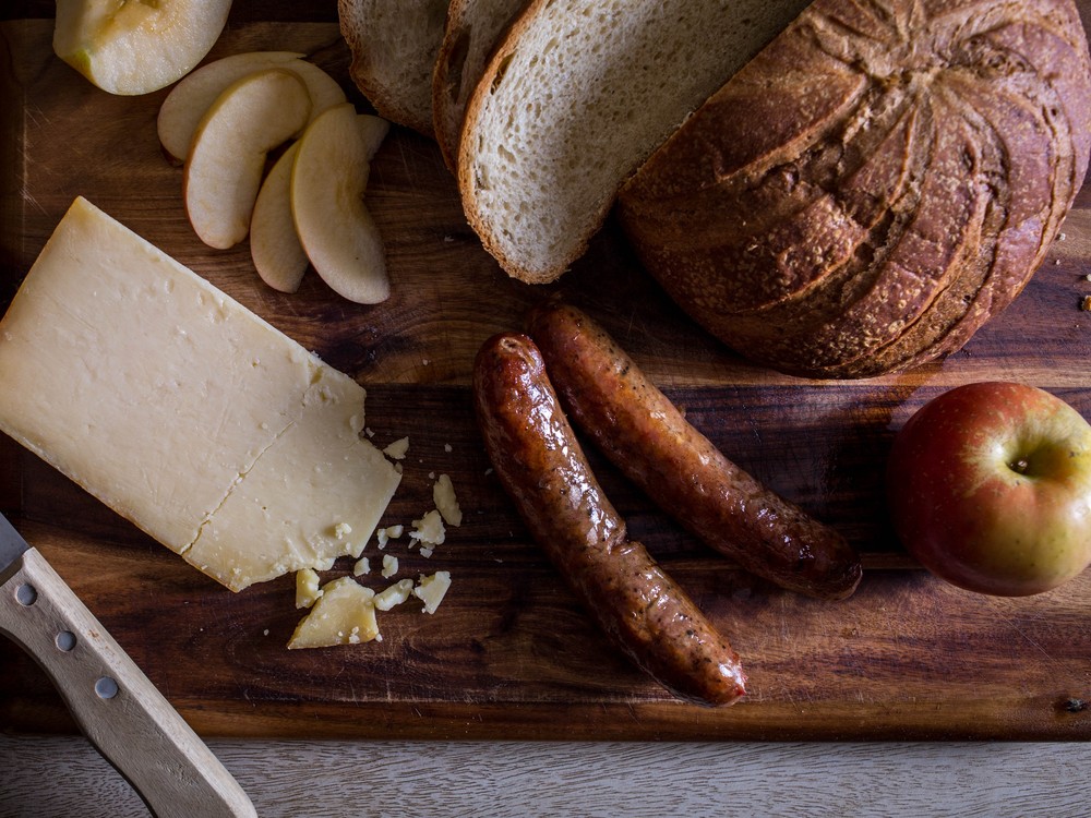 A slice of Cabot Clothbound Cheddar cheese accompanied by sausages, apples, and bread, all arranged on a wooden surface.