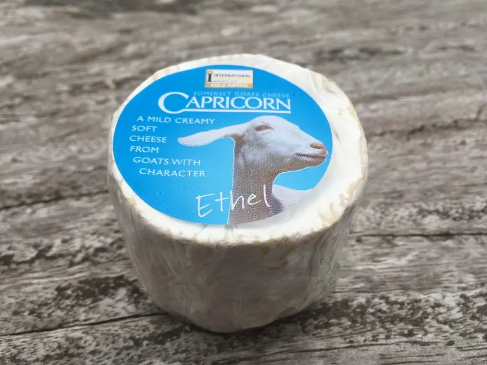 A cylindrical-shaped Capricorn Somerset Goat's Cheese, wrapped in a transparent sheet with a blue label, rests on a wooden surface