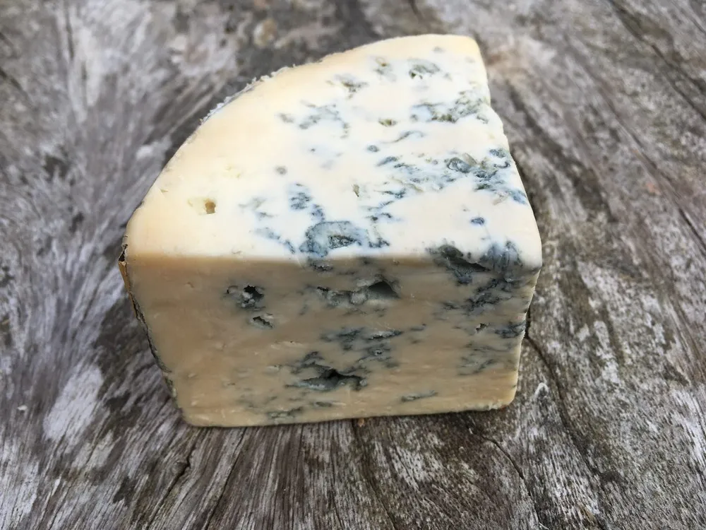 A triangular slice of Cashel Blue cheese, showcasing its creamy paste with prominent blue veins, rests on a wooden surface