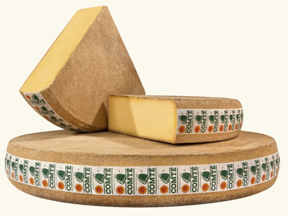 A full wheel of Comté cheese wrapped in a band cloth labeled "Comté" in green, with two chunks of the cheese resting on top. The wheel showcases the cheese's firm, dense texture and rich yellow color, highlighting its artisanal quality and French origin.