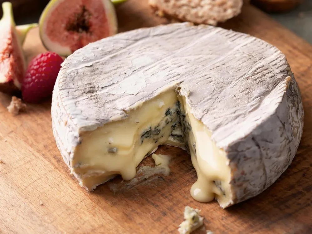 A disc of Cote Hill Blue cheese, partially sliced to reveal its creamy, runny interior with blue veins, rests on a wooden surface next to fresh figs