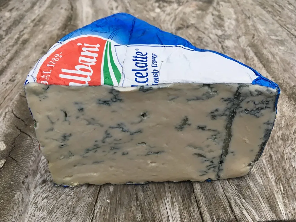 A triangular piece of Dolcelatte cheese, featuring blue veins and a crumbly interior, along with a blue wrapper, rests on a wooden surface