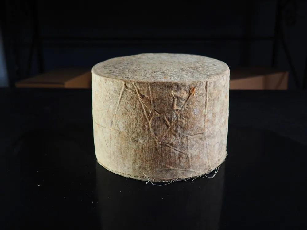 A cylindrical-shaped Traditional Ayrshire Dunlop PGI cheese, wrapped in cloth, rests on a black surface