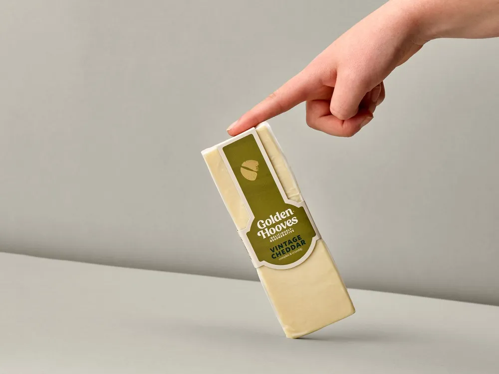 A rectangular-shaped Golden Hooves Vintage Cheddar, in its wrapper, is held by the tip of a finger against a beige background