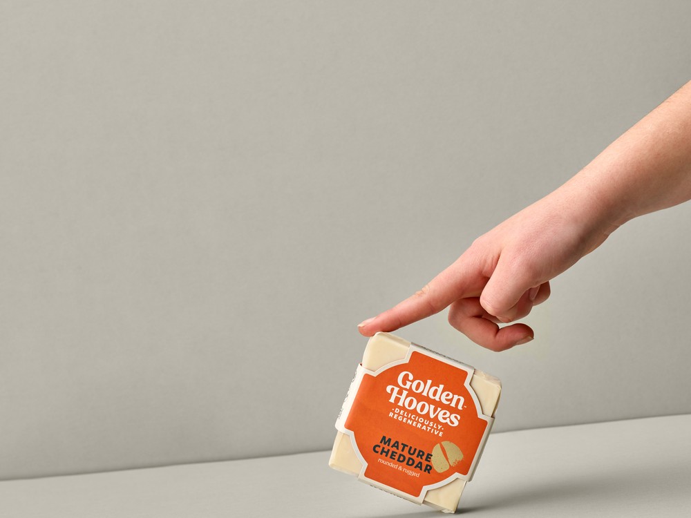 A square-shaped Golden Hooves Mature Cheddar, in its wrapper, is held by the tip of a finger against a beige background