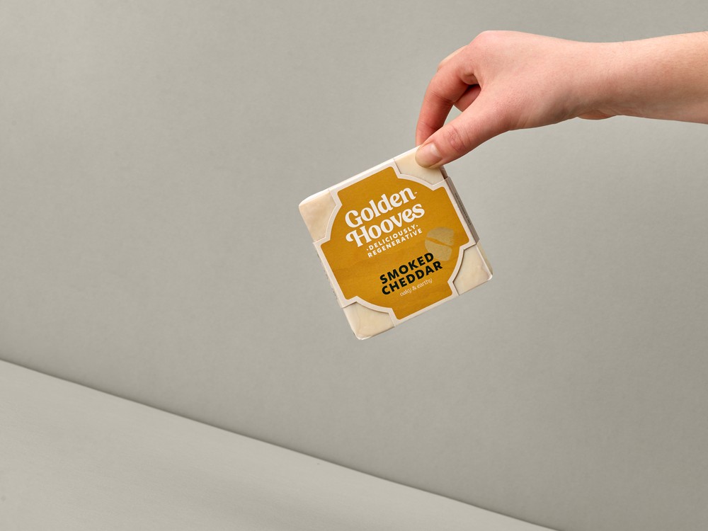 A square-shaped Golden Hooves Smoked Cheddar, in its wrapper, is held in a hand against a beige background