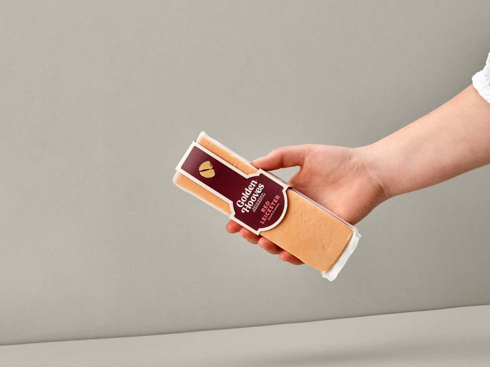 A rectangular-shaped Golden Hooves Red Leicester, in its wrapper, is held in a hand against a beige background