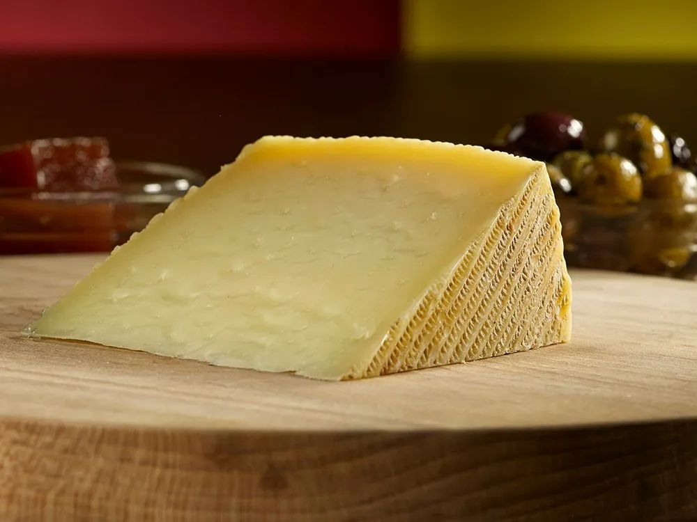 A wedge of cheese with a textured rind is placed on a wooden board, with a bowl of olives and red condiments in the background.