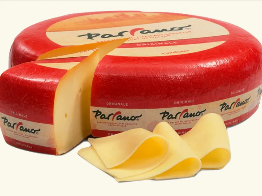 A wheel of Parrano cheese wrapped in its plastic rind is partially sliced, with thin slices arranged beside it against a white background