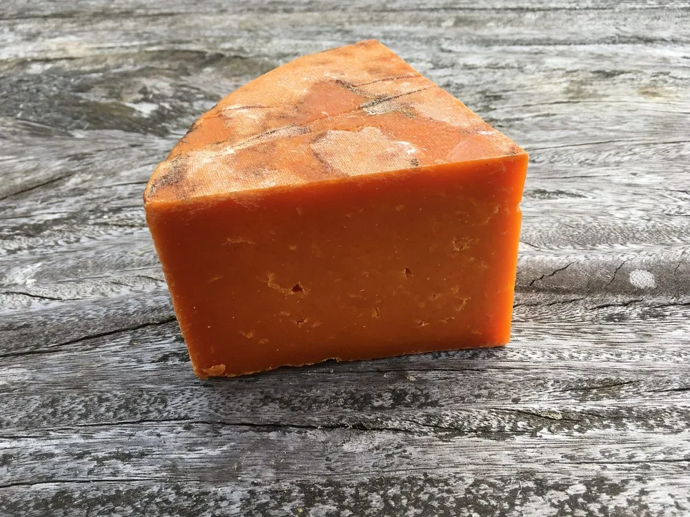 A slice of Red Leicester, displaying its vibrant orange hue from annatto, sits on a wooden surface