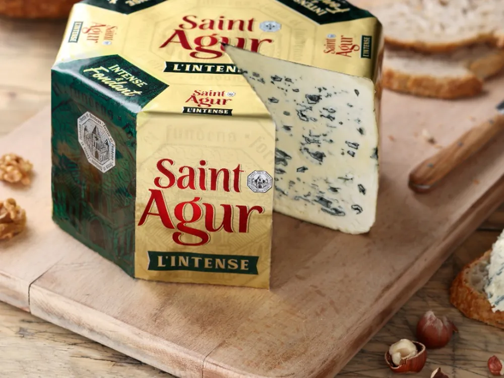 A partially cut wheel of Saint Agur cheese with its wrapper, set on a wooden board alongside a knife and some nuts