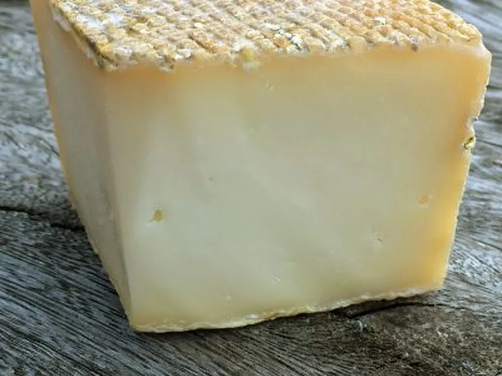 A slice of Sheep Rustler cheese, highlighting its distinctive orange-brown rind, rests on a wooden surface