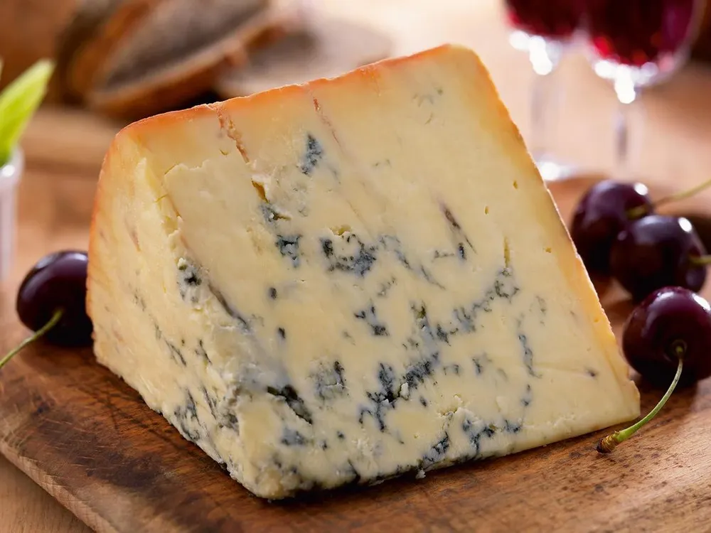 A slice of Colston Bassett Stilton, showcasing its creamy interior marbled with blue veins, is paired with fresh cherries on a wooden surface