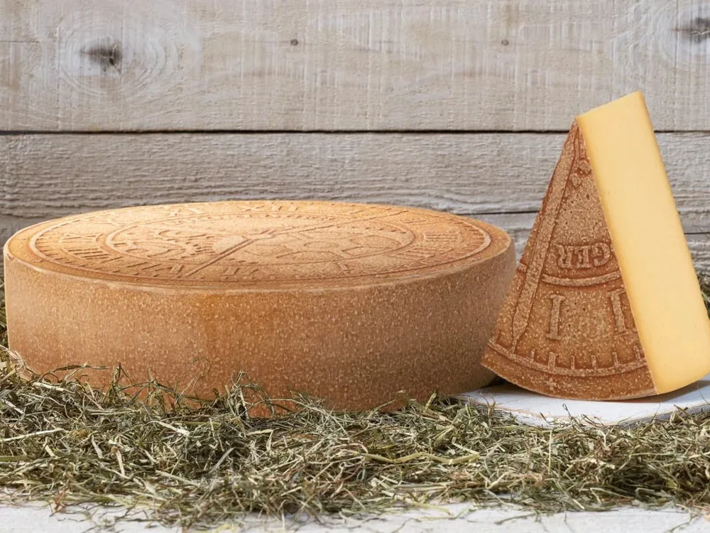 A wheel of L’Horloger cheese, accompanied by a triangular slice, rests on a white surface. Herbs are scattered around the cheese and positioned against a wooden board in the background