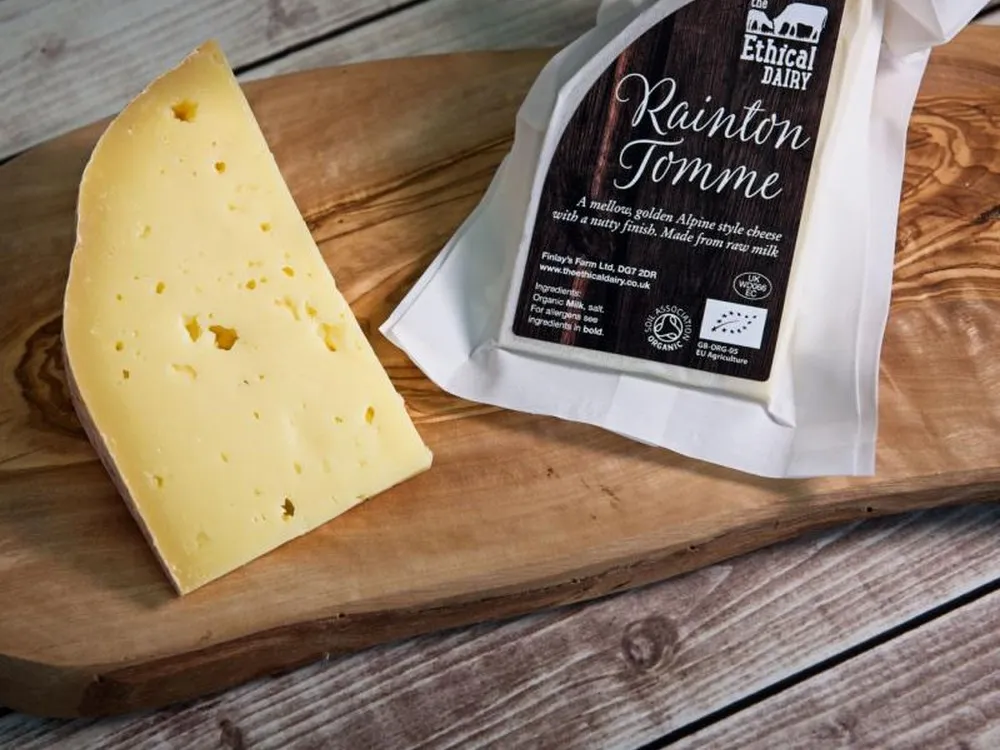 A slice of Rainton Tomme cheese, accompanied by its packaging, rests on a wooden board