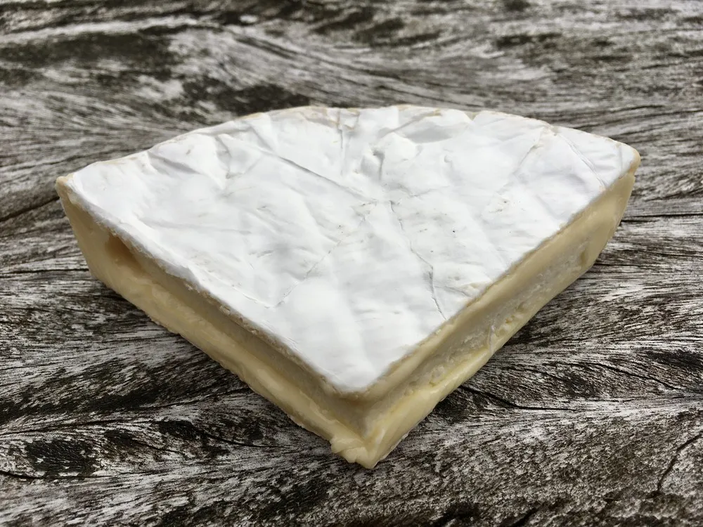 A triangular slice of Cornish Brie cheese, revealing its smooth, buttery interior, sits on a wooden board