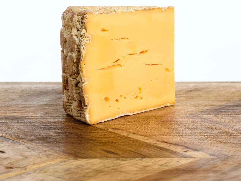 A piece of Woodside Red cheese rests on a wooden revealing orange interior and a natural rind.