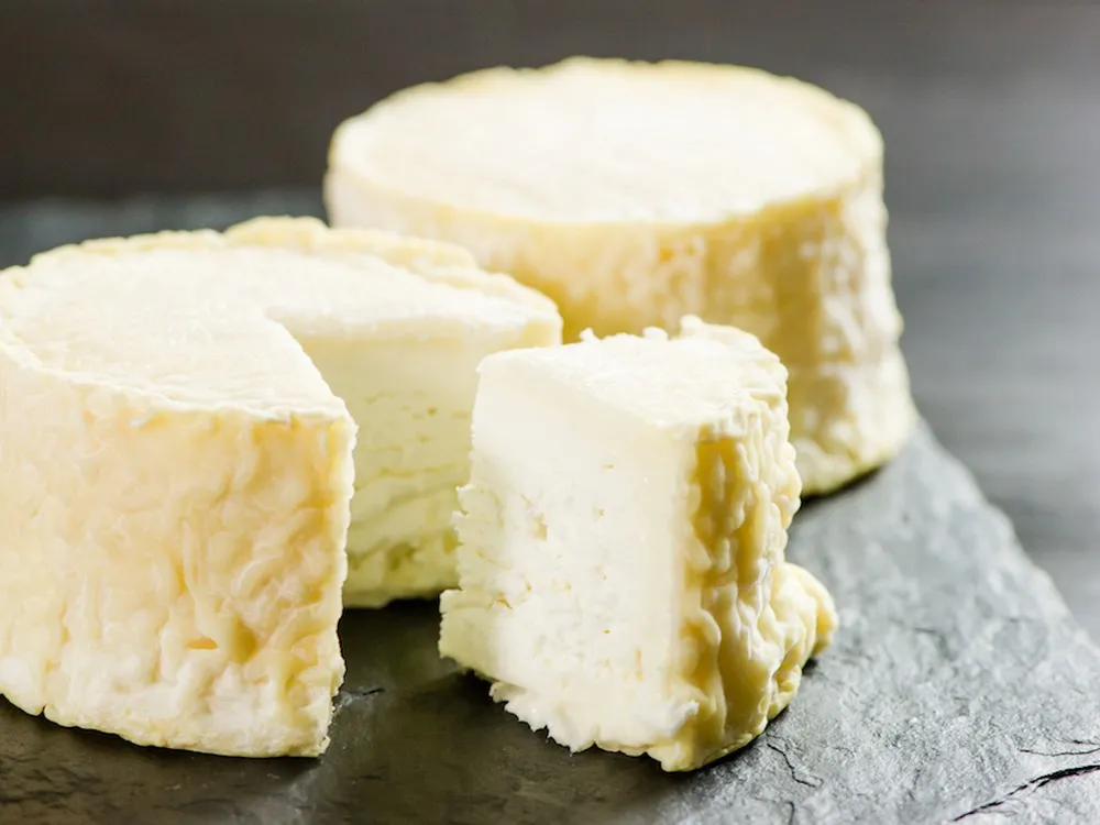 A partially cut wheel of The Manchester cheese and a whole wheel, both showing a luscious cream line beneath a thin, wrinkly rind, rest on a black surface