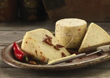 A cylindrical wheel of Northumberland Chilli cheese, with large slices revealing chili flecks, is arranged on a round wooden plate. It’s accompanied by a conical cheese sampler and red chilies, all set on a wooden surface