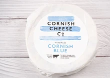 A Cornish Blue cheese wheel wrapped in white paper with a label, set against a white wooden board