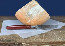 A piece of Mahón cheese, accompanied by a sharp knife, rests on butter paper placed on a wooden surface