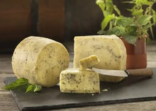 A cylindrical Northumberland Nettle cheese, along with its slices revealing a cream-colored interior speckled with herbs, is displayed on a black surface. Fresh nettle leaves and a knife on a wooden board accompany the arrangement