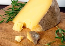 A slice of Pleasant Ridge Reserve cheese resting on a wooden board, surrounded by fresh herbs and small chunks of the cheese