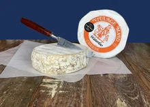 A disc-shaped Waterloo cheese is being sliced with a knife on a cutting board, next to a wheel of Waterloo cheese wrapped in white paper.