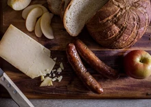 A slice of Cabot Clothbound Cheddar cheese accompanied by sausages, apples, and bread, all arranged on a wooden surface.