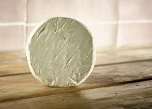 A small disc of Perl Wen cheese, featuring a bloomy white rind, stands prominently on a wooden surface