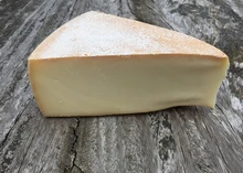 A triangular piece of Raclette cheese, featuring a brownish-orange rind and a pale yellow interior, is placed on a wooden surface