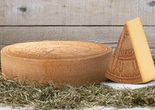 A wheel of L’Horloger cheese, accompanied by a triangular slice, rests on a white surface. Herbs are scattered around the cheese and positioned against a wooden board in the background