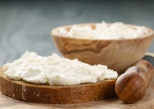 Fromage Frais spread on a slice of bread, next to a wooden bowl of cheese, with a butter knife featuring a wooden handle, all on a wooden table