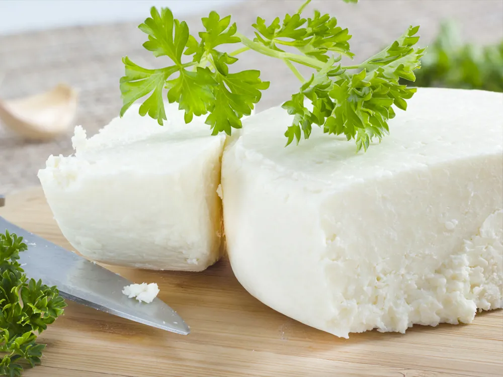 A wooden cutting board holds a block of Cotija cheese, garnished with parsley and accompanied by a knife