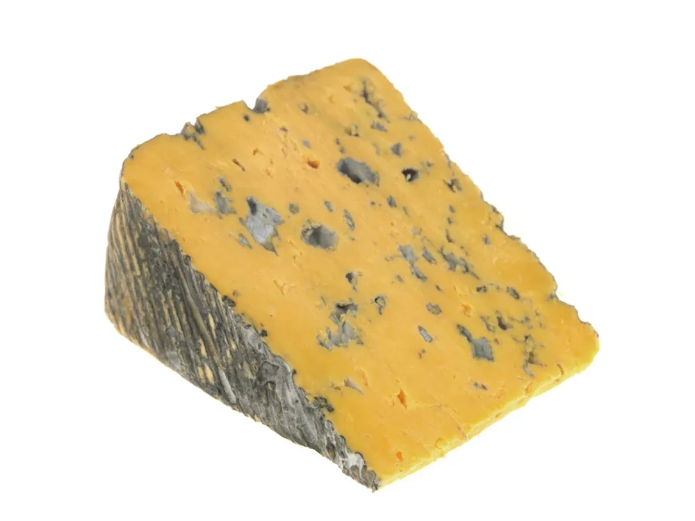 A wedge of Blacksticks Blue cheese, showcasing its soft amber-hued interior with blue veins running throughout, set against a white background.