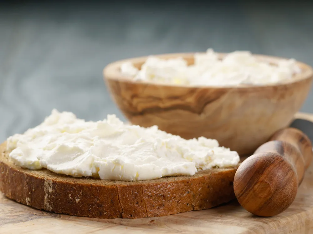 Fromage Frais spread on a slice of bread, next to a wooden bowl of cheese, with a butter knife featuring a wooden handle, all on a wooden table