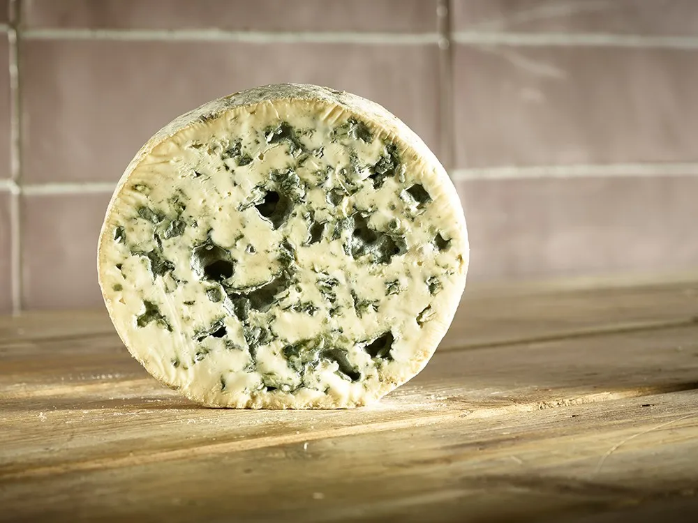 A disc of Fourme d'Ambert cheese showing it cream-coloured paste with prominent blue veins, rests on a wooden surface