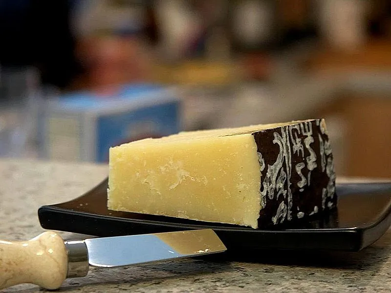 A black plate showcases a piece of Pecorino Romano cheese with its protective covering on one side, revealing its granular texture, accompanied by a knife