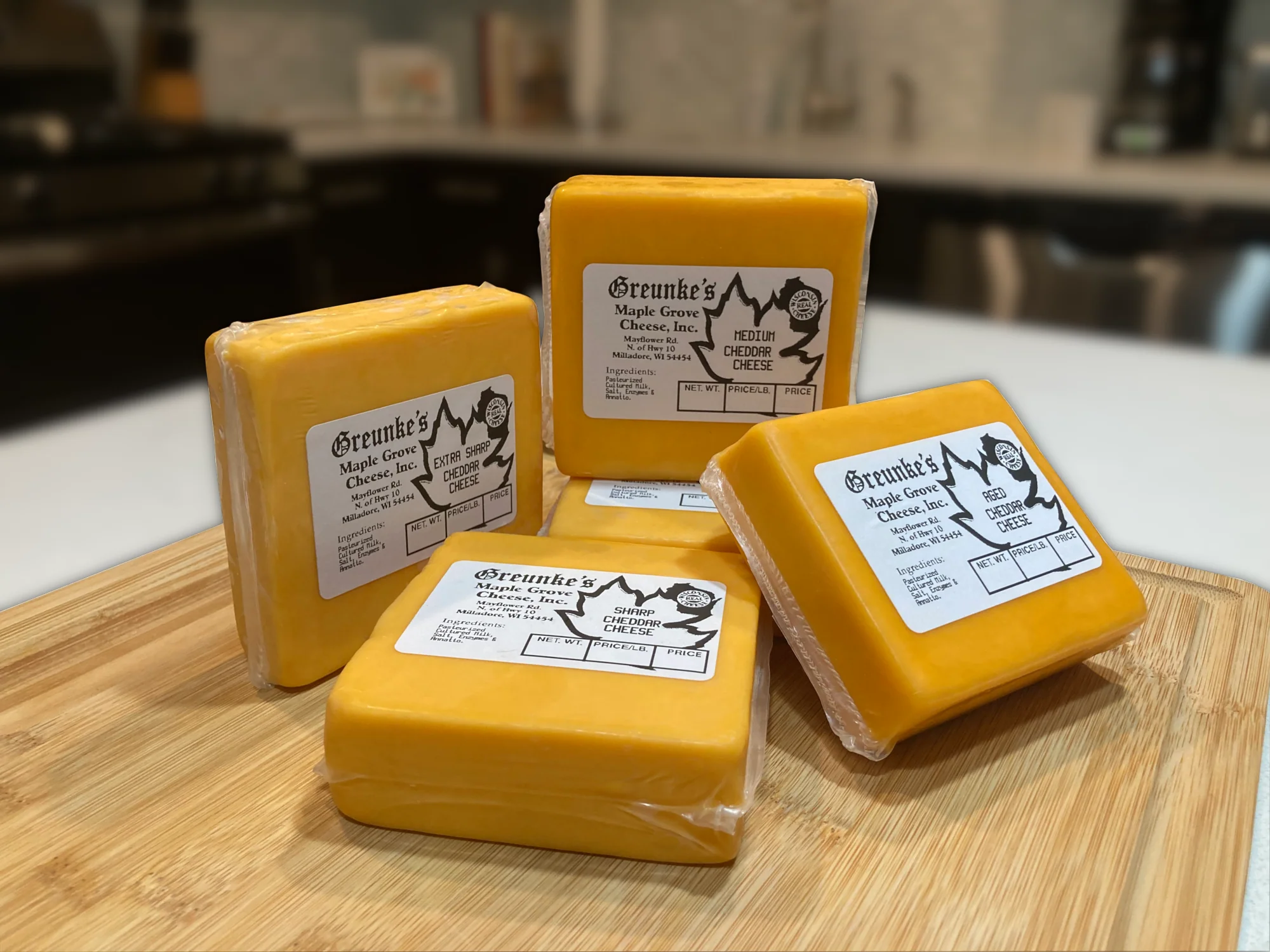 Blocks of a variety of cheddars along with Aged Cheddar, wrapped in plastic with labels on top, neatly arranged on a wooden table.