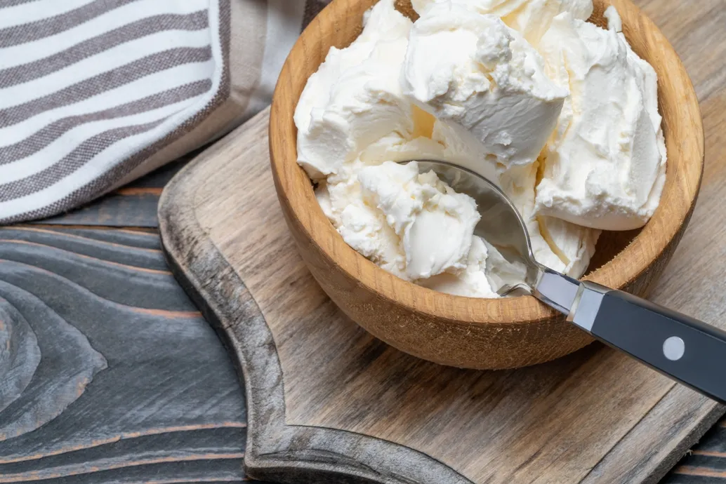 A wooden bowl filled with scoops of Mascarpone Cheese, placed on a wooden cutting board. A metal spoon with a black handle rests inside the bowl. A striped cloth napkin is partially visible in the background.