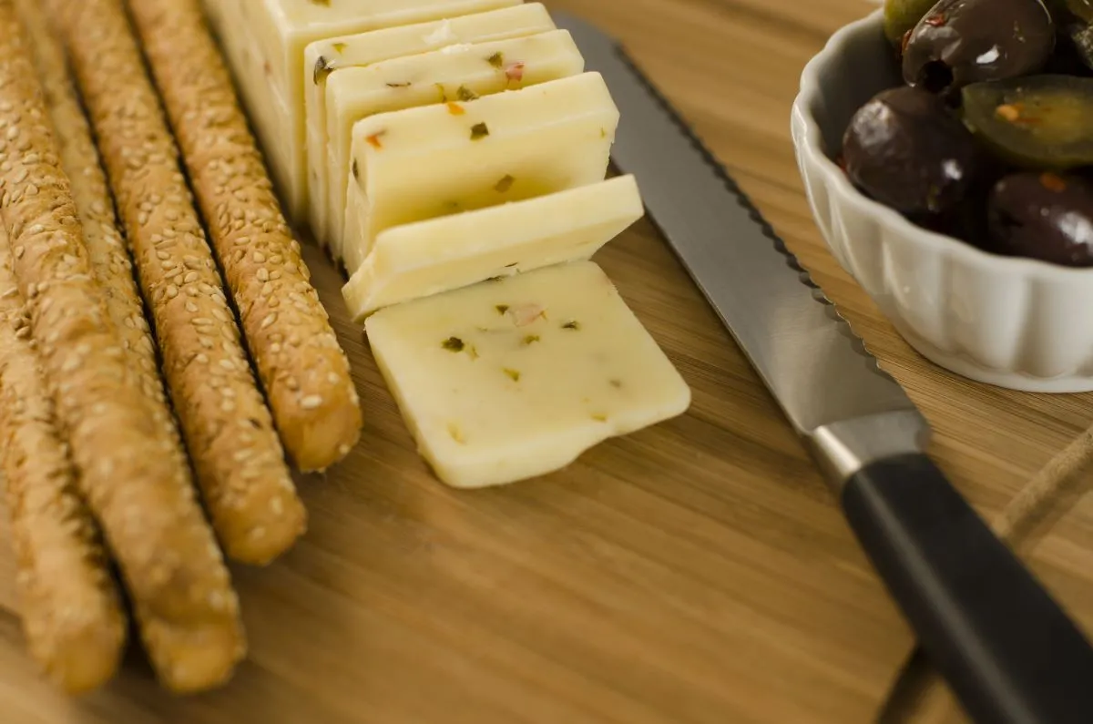 Sliced Pepper Jack cheese with sesame seed breadsticks, Mediterranean olives, and hot peppers on a wooden cheese board, with a knife