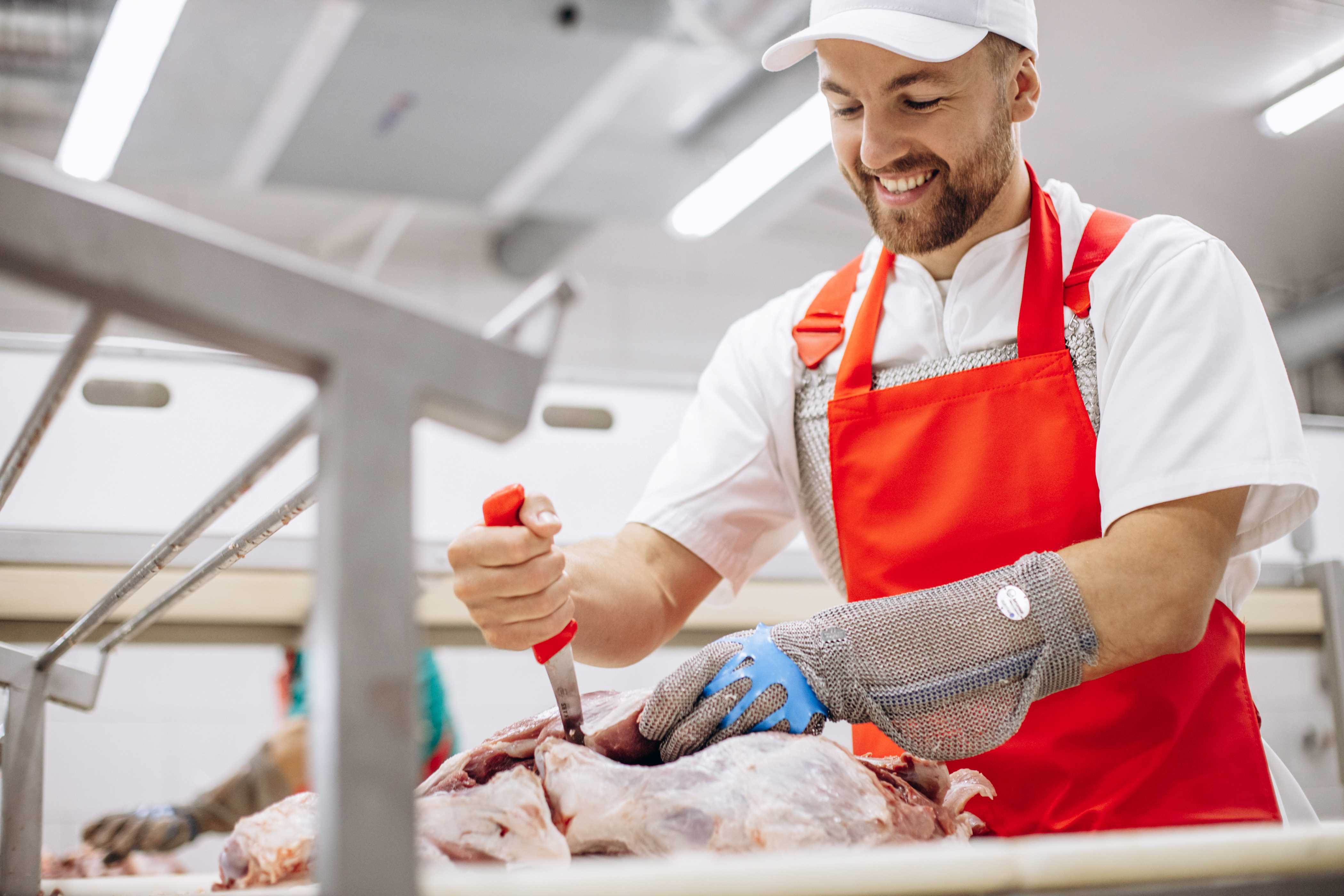 A butcher with a red apron cutting a piece of meat with a big knife